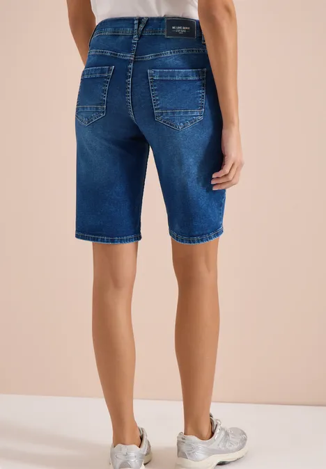 Achteraanzicht van een persoon in donkerblauwe denim bermuda en witte sneakers voor een beige achtergrond.