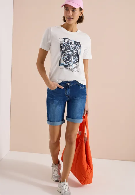 Vrouw in wit T-shirt met print, spijkerbroekshort, sneakers en rode tas.