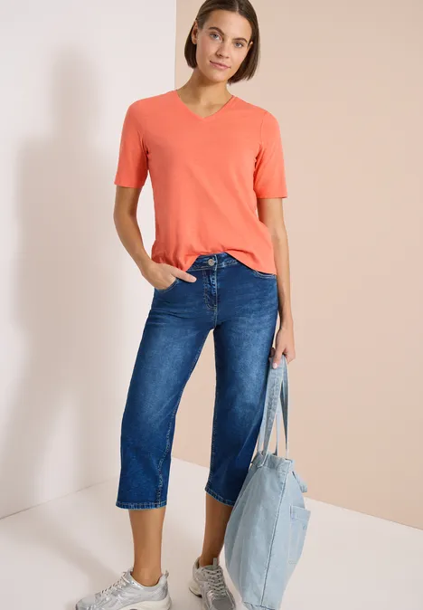 Femme en T-shirt corail col V et jean court bleu, tenant un sac en denim clair sur fond neutre.