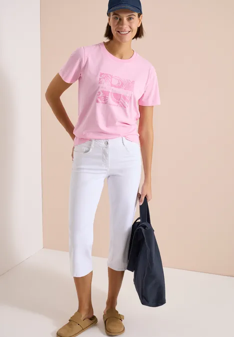 Frau trägt rosa T-Shirt, weiße Capri-Jeans, braune Slipper und hält eine dunkelblaue Stofftasche.