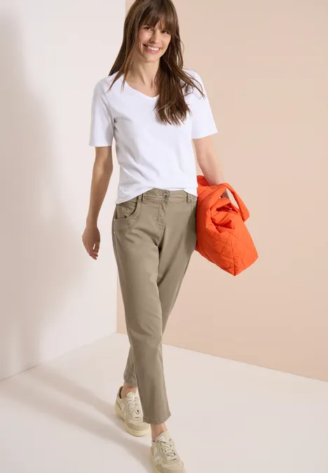 Frau in weißem T-Shirt, khakifarbene Hose und weißen Sneakern, trägt eine orangefarbene Tasche vor hellem Hintergrund.