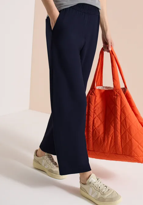 Persoon draagt marineblauwe wijde broek en sneakers, houdt een oranje gewatteerde draagtas vast.