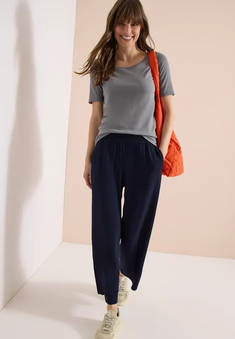 Een vrouw draagt een grijs t-shirt, een donkerblauwe broek, witte sneakers en een oranje schoudertas.