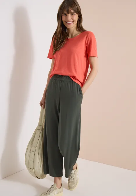 Vrouw in koraalkleurig T-shirt en wijde donkergroene ballonbroek, met een beige draagtas.