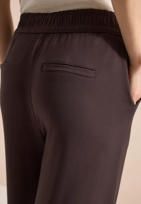 Nahaufnahme: Person trägt dunkelbraune Hose mit elastischem Bund, Seitentaschen und Paspeltasche.
