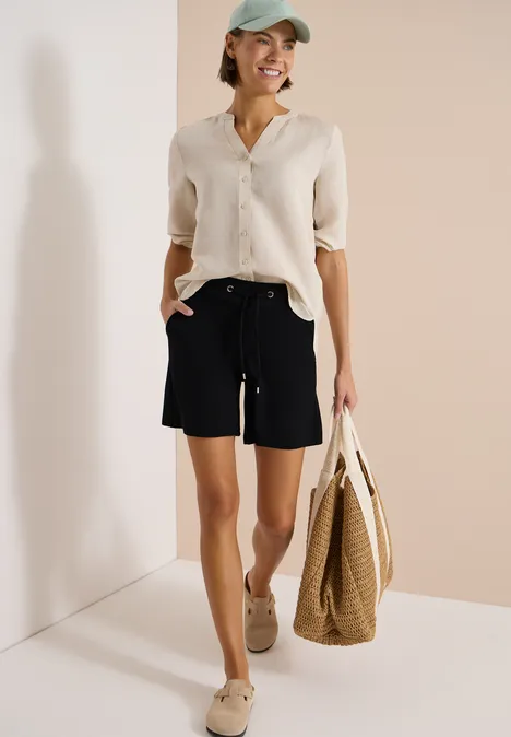 Vrouw in witte blouse, zwarte shorts en beige sandalen met grote rieten tas tegen een lichte achtergrond.