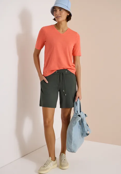 Persoon draagt een koraalkleurig T-shirt, donkergroene short, sneakers en houdt een spijkerjasje.