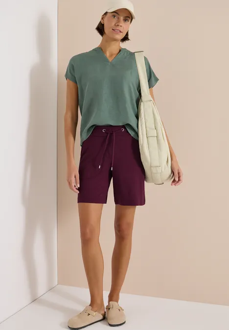 Persoon met groen shirt, paarse shorts, sandalen en schoudertas voor een lichte achtergrond.