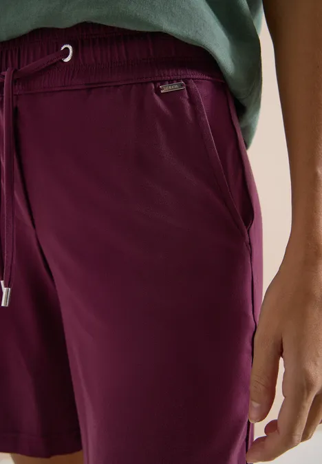 Close-up van een persoon in bordeauxrode shorts en groen shirt, met zak en trekkoord.