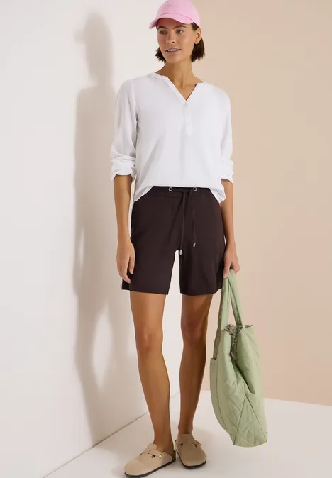 Vrouw in wit hemd en zwarte short met beige instappers, voor een lichte muur met groene gewatteerde tas.