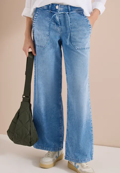 Personne porte un jean large bleu clair et des baskets blanches, tenant un sac vert foncé.