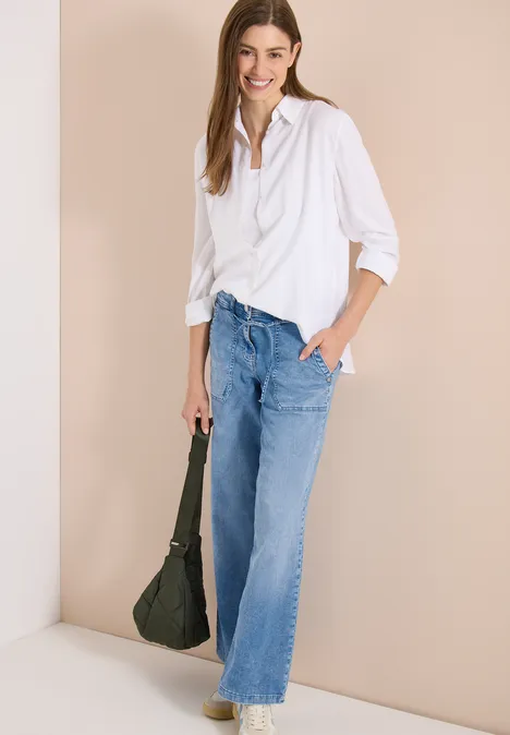 Femme en chemise blanche et jean ample bleu, pose décontractée, tient un sac sombre sur fond neutre.