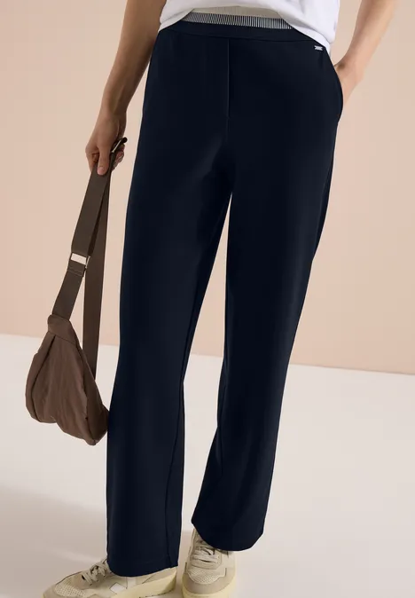 Dunkelblaue weite Hose, weiße Sneaker und braune Tasche in lässiger Pose.