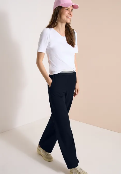 Frau in weißem T-Shirt, schwarzer Jogginghose, Sneakern und rosa Cap vor hellem Hintergrund.