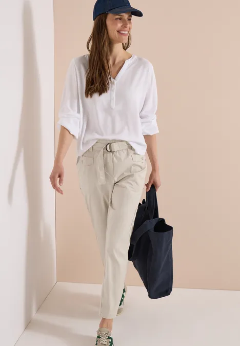 Frau in weißer Bluse, beiger Hose und dunkler Cap trägt eine schwarze Tasche.
