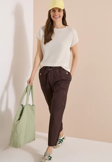 Frau in weißem T-Shirt, braunen Hosen, grüner Tasche und passenden grünen Sneakern vor beigem Hintergrund.