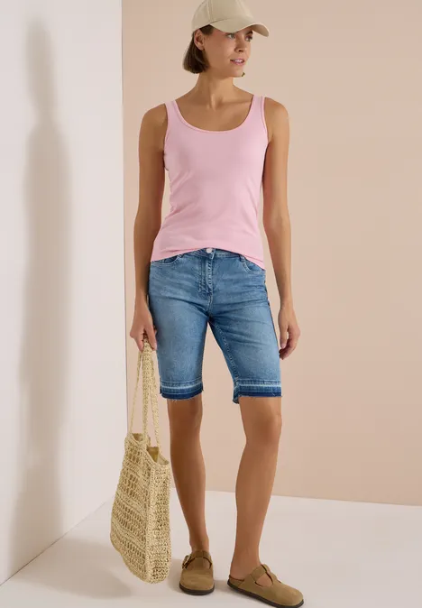 High Waist Wide Leg Jeans im Loose Fit light blue washed Frau in rosa Tanktop, Jeans-Bermuda, Baseballcap, trägt Strohkorbtasche und bequeme Loafers vor beige Wand.
