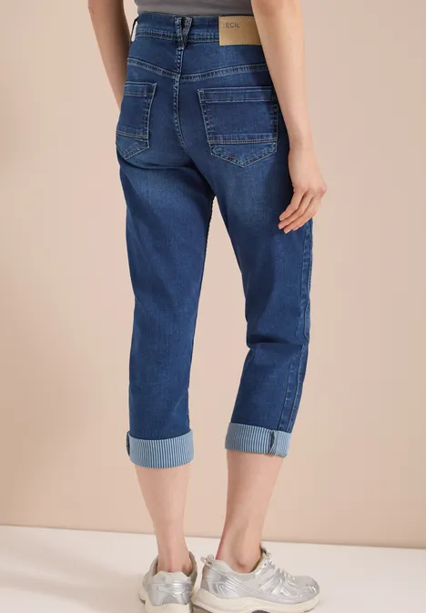 Mid Waist straight leg jeans in casual fit mid blue wash Persoon van achteren tegen beige achtergrond, met opgerolde blauwe jeans en witte sneakers.