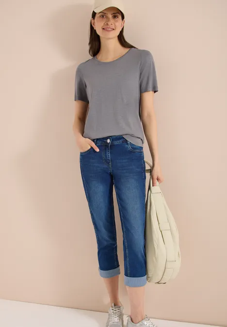 Mid Waist straight leg jeans in casual fit mid blue wash Een persoon draagt een grijs T-shirt, omgeslagen jeans, glinsterende instappers en houdt een lichte tas vast.