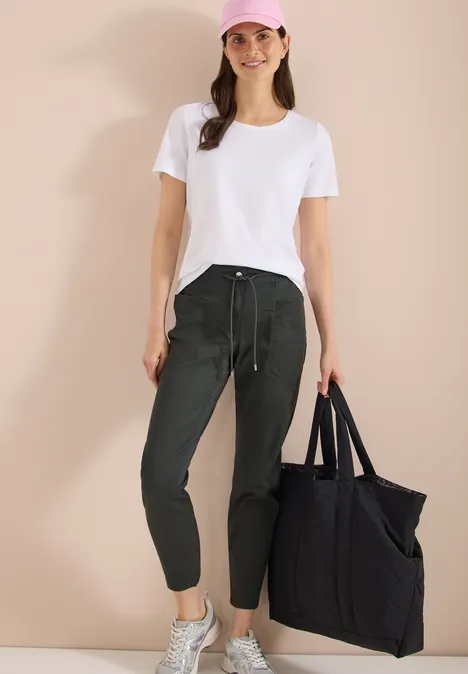 Mid Waist Slim Legs broek in een gewassen look clear khaki Vrouw in wit T-shirt, groene joggingbroek, witte sneakers en grote zwarte tas tegen beige achtergrond.