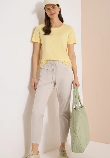 Frau in gelbem T-Shirt, hellgrauen Hosen und grünem Stepp-Shopper vor schlichtem, pastellfarbenem Hintergrund.
