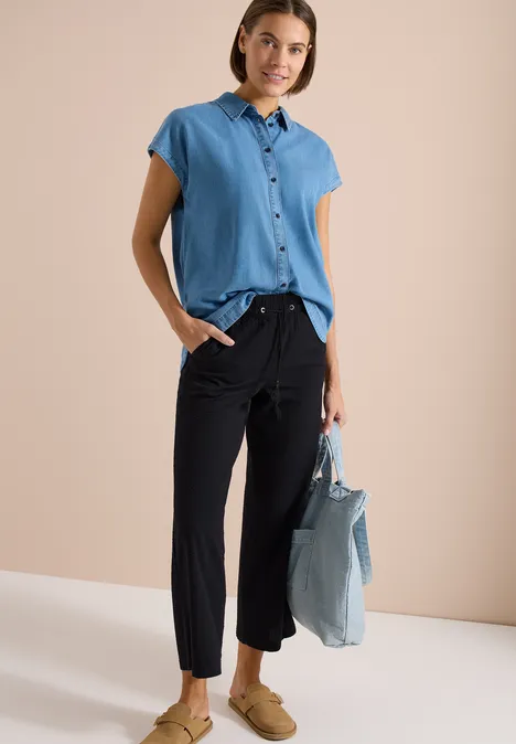 Persoon draagt lichtblauw overhemd met korte mouwen, zwarte broek, beige loafers en een lichtblauwe tas.