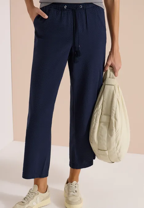 Loszittende marineblauwe broek met aantrekkoord, gecombineerd met lichte sneakers en een lichte jas in de hand.