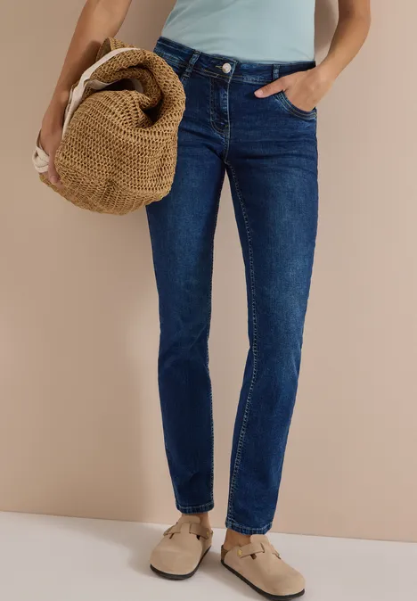 Mid Waist Straight Leg Jeans im Casual Fit mid blue used wash Schlanke blaue Jeans, beigefarbene Slipper und geflochtene Tasche vor beigem Hintergrund.