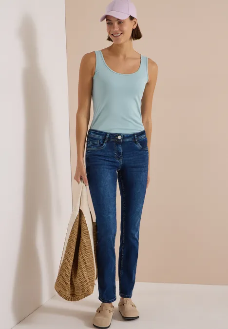 Mid Waist Straight Leg Jeans im Casual Fit mid blue used wash Junge Frau in Jeans, ärmellosem Top und Cap, trägt eine geflochtene Tasche.