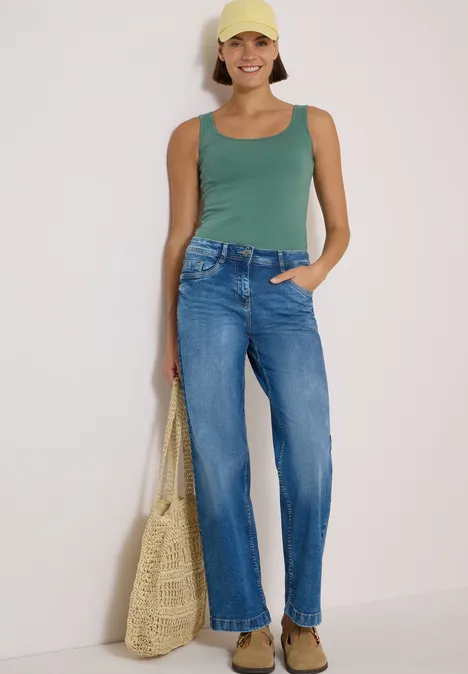 High Waist Wide Leg Jeans im Loose Fit mid blue wash Frau mit gelber Cap, grünem Top, blauen Jeans und Strohtasche lächelnd