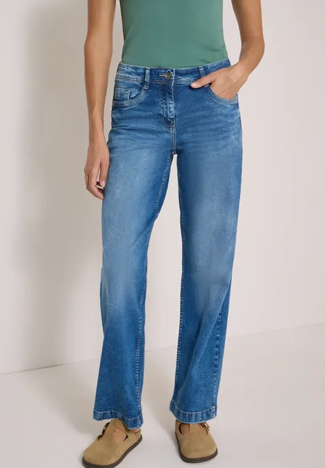 High Waist Wide Leg Jeans im Loose Fit mid blue wash Stehende Person mit hellblauer, gerader Jeans im Used-Look und beigefarbenen Wildleder-Slippern.