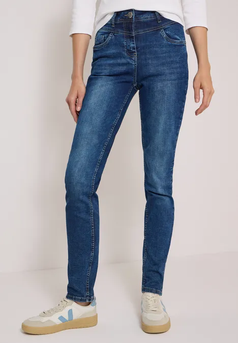 Persoon draagt blauwe skinny jeans en witte sneakers, minimalistische achtergrond.