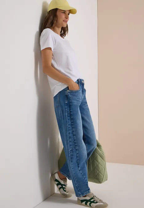 Frau lehnt locker an Wand, trägt weißes Shirt, weite Jeans, gelbe Mütze, grüne Sneaker und hält gesteppte Jacke.