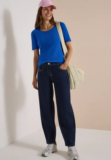 Frau mit rosa Cap, blauem Shirt, weiten Jeans, silbernen Sneakern und Tasche vor beigem Hintergrund.