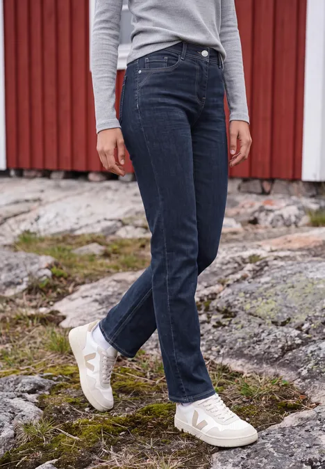 Frau in weißen Sneakern und dunklen Jeans steht auf felsigem, moosbedecktem Boden vor roter Holzwand.
