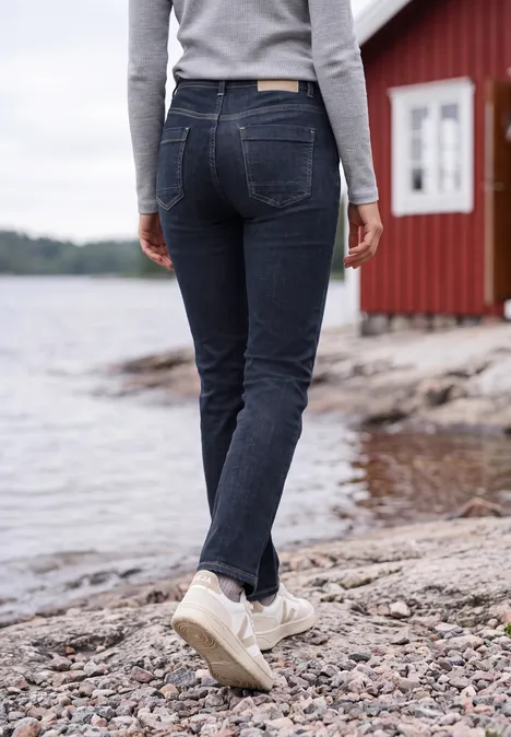 Frau im grauen Pullover und Jeans läuft in weißen Sneakern über einen Steinstrand neben einem roten Haus.