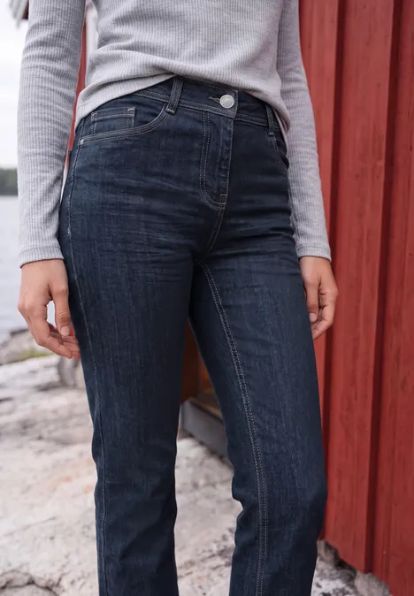 Eine Person trägt dunkle Jeans und graues Langarmshirt und steht vor einer roten Holzwand.