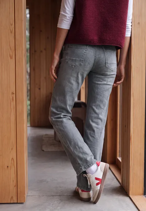 Person von hinten in grauen Jeans und weißen Sneakers steht in einer hölzernen Türöffnung.