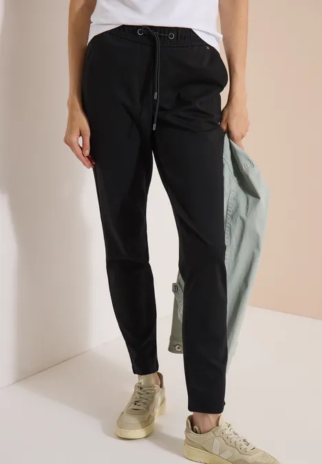 Persoon draagt een zwarte joggingbroek met trekkoord, sneakers en houdt een groene jas over de schouder.
