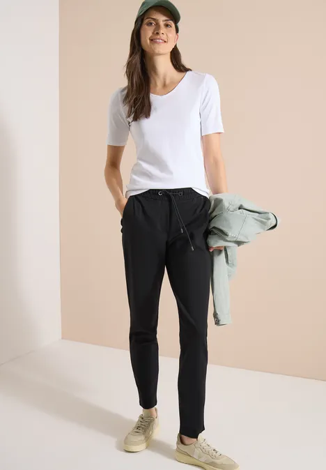Vrouw met wit T-shirt en zwarte broek houdt een jas vast, minimalistische achtergrond, draagt sneakers.