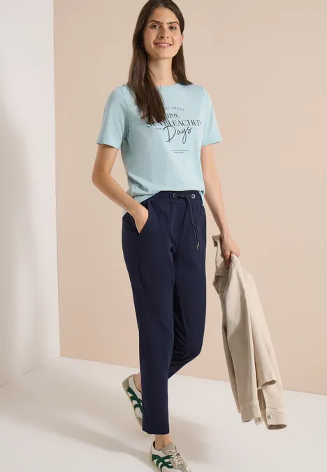 Vrouw in turquoise t-shirt, donkerblau broek en witte sneakers, met een beige blazer in de hand.
