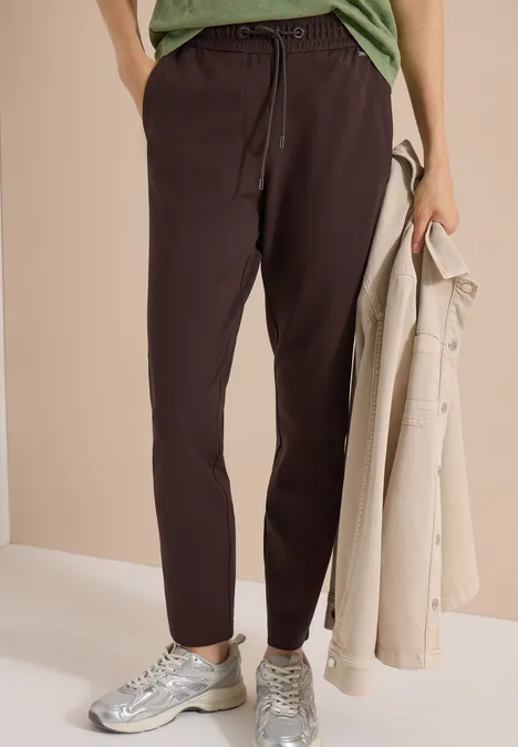 Personne porte un pantalon marron foncé, baskets argentées et tient un manteau beige sur fond neutre.
