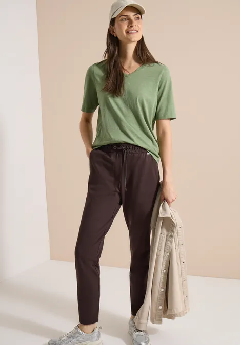 Femme casquette, t-shirt vert et pantalon noir, tenant une veste beige à la main.