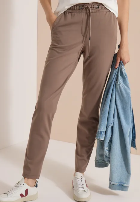 Femme en pantalon de jogging marron, baskets blanches, tenant une veste en jean; look décontracté.