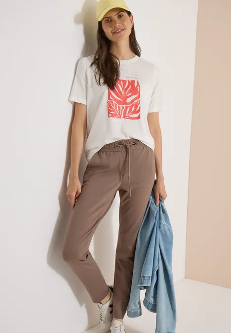 Femme en t-shirt blanc à motif rouge et pantalon beige, tenant une veste en jean devant un fond clair.