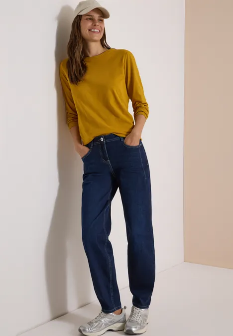 Eine lächelnde Frau lehnt an einer Wand, trägt gelbes Shirt, Jeans und weiße Sneakers.