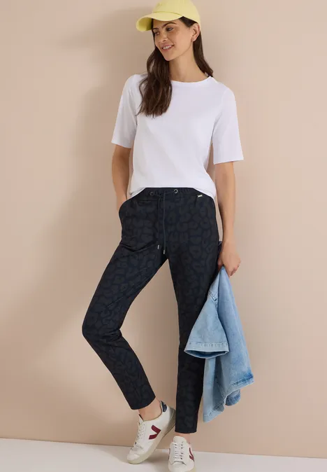 Mid Waist Slim Leg Hose im Casual Fit dark navy blue Frau trägt weisses T-Shirt, gemusterte Dunkelhose, weisse Sneaker, gelbe Kappe, hält Jeansjacke.