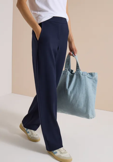 Person trägt dunkelblaue, weite Hose, weiße Sneakers und hält eine hellblaue Denim-Tasche.