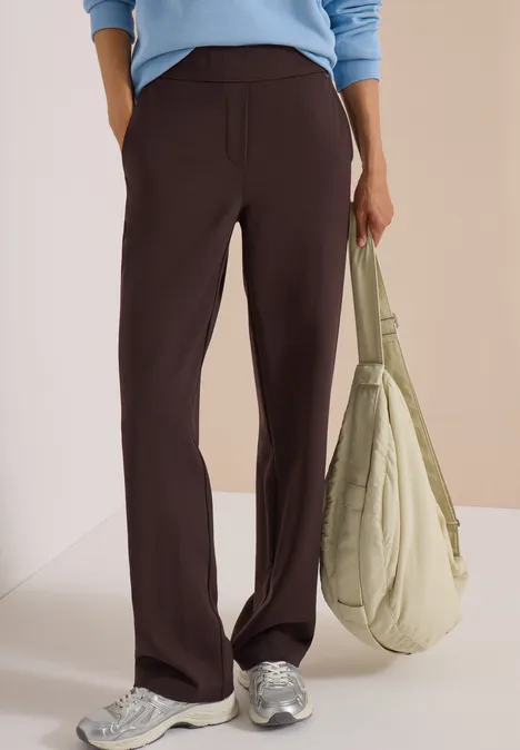 Person trägt weite, dunkelbraune Hose, hellblaues Oberteil, weiße Sneaker und hält eine hellgrüne Tasche.