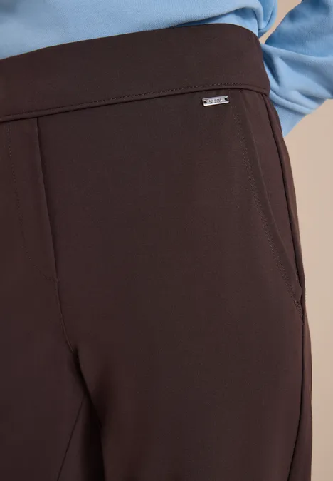 Auf dem Bild: dunkle, elegant geschnittene Hose mit kleinem silbernen Logo am Bund, kombiniert mit blauem Oberteil.